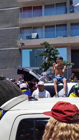 The video shows a crowd of people celebrating in the street. Most people are surrounding white cars and holding Venezuelan flags. One man is sitting on the roof of a car without a shirt. The scene takes place in a city, with apartment buildings visible in the background. The weather appears sunny. The crowd is densely packed, and a sense of celebration prevails. The sounds are loud music and cheering.