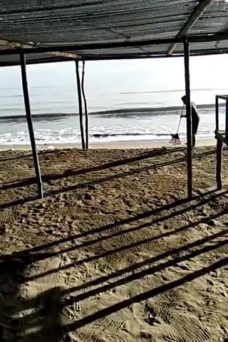 A person stands on a beach and sweeps with a rake. The person is silhouetted and indistinct. The beach is sandy and has many footprints. A beach shade structure creates shadows in the foreground. The sea is visible behind the person, with gentle waves lapping the shore. The sky is overcast.
