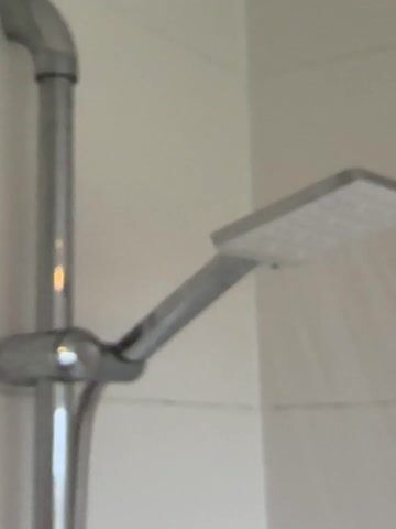 A shower head is visible in the frame. It is a square, chrome-colored shower head with water flowing out of it. The shower head is attached to a vertical chrome pipe. The background consists of white tiled walls.