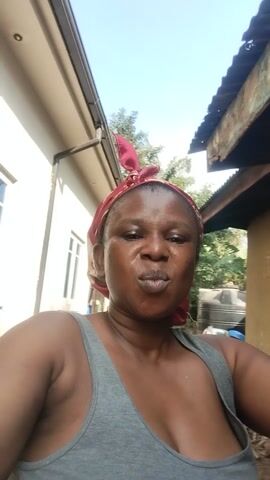 A woman with a red and white patterned headscarf and a grey tank top poses for the camera in Abor, Nigeria. She stands outdoors, with a building featuring a light brown wall and a corrugated metal roof visible behind her. The scene is brightly lit, suggesting daytime. She makes various playful facial expressions, first puckering her lips, then opening her mouth wide in a smile or laugh while extending her left hand with her palm facing up. Some unclear, scribbled text is visible on the wall behind her.