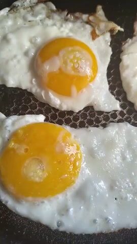 Two fried eggs are being cooked in a dark, textured pan. The eggs are sunny-side up, with bright yellow yolks and translucent white. The whites are starting to crisp and brown slightly at the edges. Bubbles are visible in the whites, indicating active cooking. The camera is positioned directly above the pan, offering a close-up view of the eggs. The lighting is bright, highlighting the glistening yolks and whites.