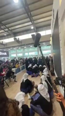 Certainly! Here's a description of the video:

The video captures a group of people performing a traditional Middle Eastern musical performance. They are sitting in a circle on a wooden floor in what appears to be a large exhibition hall or indoor space. They are wearing traditional clothing, consisting of dark blue or black outfits with white head coverings. Some are playing small hand drums or other percussion instruments. 

A crowd of people stands around the performance area, watching and recording the event with their phones and cameras. A professional microphone and camera are positioned close to the group, capturing audio and video. The surroundings are brightly lit, with a high ceiling and modern architectural elements visible. There are also booths and displays in the background, suggesting that this is taking place at an exhibition or fair.