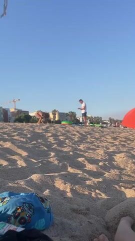 A low-angle shot captures a sandy beach under a clear blue sky. In the foreground, a person's legs and feet are visible, with some fabric of clothing lying on the sand. In the middle ground, a person on their back with their legs in the air is visible. To the right, a person is standing and looking at their phone. Further back, an orange umbrella provides shade for a person or people sitting underneath. Several other beachgoers, along with beach chairs and inflatable toys, are scattered across the sand in the distance. Palm trees and buildings are visible on the horizon. The overall scene depicts a sunny day at a crowded beach.