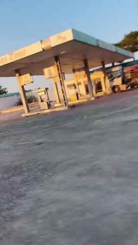 The video pans across a gas station and an adjacent building under a clear sky during daylight. The gas station has a covered canopy with "Haci Zaur" written on it, and two "DIZEL" fuel pumps. A small building with large windows is visible to the right of the gas station. Further to the right, a sign with a map of a country and text is seen, with a small green bush in front of it. The ground is paved with gravel and dirt. The scene appears to be in a rural or semi-rural area.