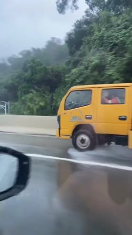 The video shows a person driving on a highway in the rain. The camera is inside the vehicle, looking out the front windshield. The road is wet and there are many water droplets on the windshield. Trees and hills are visible on the side of the road. A yellow truck is seen driving in the opposite direction. The person driving the car is talking and appears to be in distress. The video is shaky and disorienting, likely due to the poor road conditions and the driver's emotional state.