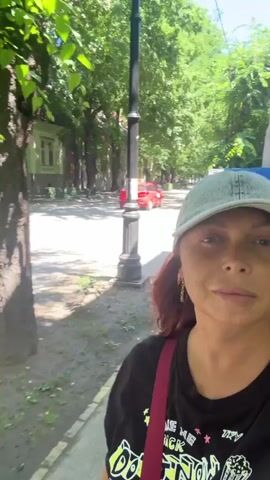 A woman wearing a light blue baseball cap and a black t-shirt with a graphic print is walking outdoors on a sidewalk next to a street lined with trees. She is holding her phone to record a selfie video. She has reddish-brown hair and is wearing earrings and a red strap across her chest from a bag. The street is paved, and there are houses and parked cars visible across the street. The trees have dense green foliage, and the sun is shining, casting shadows on the ground.