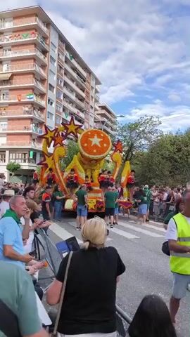 A vertical, full-shot video captures a parade float adorned with yellow and orange flowers and stars, shaped like a giant orange. People in green shirts and blue shorts stand on and around the float. A crowd of onlookers, many wearing green scarves, watches from the side of a street lined with a tall brick building. The sky is blue with white clouds. The float is moving slowly down the street.