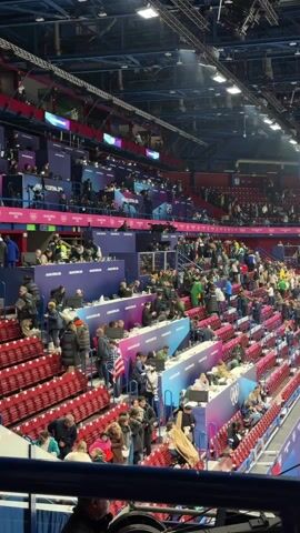 The video shows a view inside a large arena filled with people in seats. The seats are mostly red, with some blue areas designated for VIPs or media personnel. Many people are standing and moving around, while others are seated. There's an ice rink in the center of the arena. Overhead, there are various platforms occupied by people, possibly media or event staff. The ceiling is high, with a complex network of lighting fixtures and structural elements. The atmosphere seems active and energetic, suggesting a break or transition period during an event.