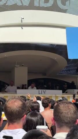 A crowd of people stands below a white ledge with a “LIV BEACH” sign, facing a small group of people on the ledge. A man in a white t-shirt is at the center of the group on the ledge. Some people wear hats, others are wearing sunglasses and the crowd is surrounded by others, some of whom are shirtless. The location seems to be outdoors.