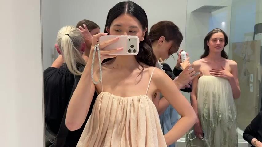 The scene is a crowded room, likely a dressing room or backstage area, as indicated by the presence of several people and wardrobe items. The central figure is a young Asian woman, dressed in a light-colored, sleeveless dress. She is standing in front of a large mirror, holding up a pink iPhone to take a selfie. The phone has a silver chain attached to it.  Behind her, there are at least five other people. One woman in a white dress is standing in front of a shower unit. Another person is fixing her hair and another is holding a phone. The background is white and brightly lit, with a minimalist aesthetic. The overall atmosphere seems busy and focused on preparation.
