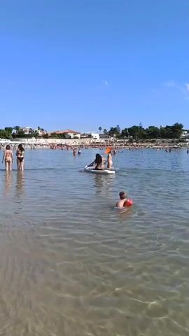 This is a low-angle, wide-shot video of a crowded beach on a sunny day. The water is clear and shallow, with gentle waves. In the foreground, two individuals are partially submerged in the water, with one wearing an orange flotation device around their waist. Further out, a person is paddleboarding, and another person is sitting on the paddleboard behind them. The background is filled with many people swimming and wading in the water. Along the shoreline, there are buildings with terracotta roofs and trees. The sky is a clear, bright blue.