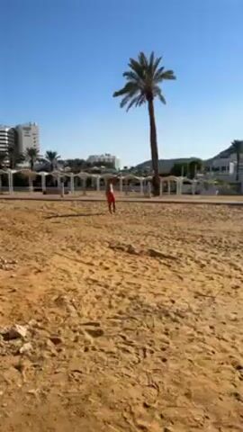 A video shows a man playing tennis on a sandy beach on a sunny day. The man is wearing orange shorts and a hat. He hits a tennis ball back and forth. Behind him are beach umbrellas, palm trees, buildings, and mountains. The beach is empty except for the man.