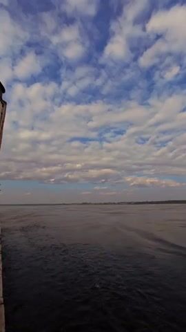 The video shows a wide shot of a dark body of water. The sky above is filled with white and grey clouds against a blue background. The horizon is flat and appears to be land, possibly with some trees or buildings visible in the distance. There are small waves on the water, creating a textured surface. The camera is moving, as indicated by the slight blur and the perspective shift, suggesting it is being filmed from a moving vessel.