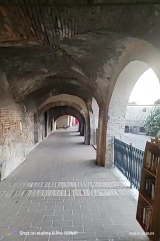 This video shows a first-person perspective walking through a stone corridor with arches. The corridor is paved with bricks and has stone walls on the left. On the right, there is a black metal fence and beyond that, a courtyard with a building and some trees. The ceiling of the corridor is arched and made of stone and brick. The video is dated July 30, 2026. A person in red is briefly visible at the end of the corridor in the distance.