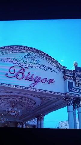 The video begins with a shot of a sign that reads "Bisyor". The sign is ornate with decorative elements and a cursive font. The scene then transitions to an indoor setting with white tiled walls. In the center of the frame is a large, circular oven with a black interior. To the right of the oven, there is a metal stand holding a large metal bowl filled with pieces of meat. A young man wearing a white shirt and a maroon apron is standing in front of a chopping board. He is holding a cleaver and appears to be in the process of preparing the meat. He is looking down at the meat and the cleaver, focused on his task. The oven appears to be a tandoor, commonly used for cooking in Central Asian cuisine. The overall impression is a food preparation area, likely in a restaurant or commercial kitchen.