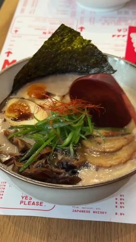 A close-up shows a bowl of ramen being stirred with a large, wooden spoon. The ramen contains noodles, sliced pork, mushrooms, green onions, half of a soft-boiled egg, chili strings, and a sheet of seaweed. The broth is a milky color. The bowl sits on a table with a menu beneath it, displaying items like “Japanese whisky” and prices. The spoon scoops up broth and noodles, revealing more of the ramen’s contents.