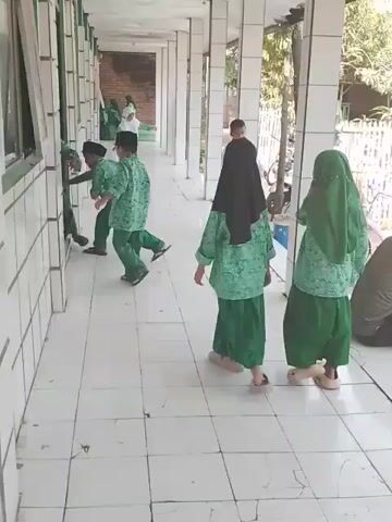 The video shows a group of children in a school hallway. The children are wearing green patterned shirts and either green pants or green skirts, depending on their gender. Some of the girls are wearing hijabs, one black and one green. The hallway has white tiled floors and white walls with a green trim. The hallway is covered, with pillars providing support. Some of the children are lined up against the wall near a window, while others are standing or walking in the hallway. A bicycle is visible in the background. The children appear to be engaged in an activity or game. The camera is stationary.