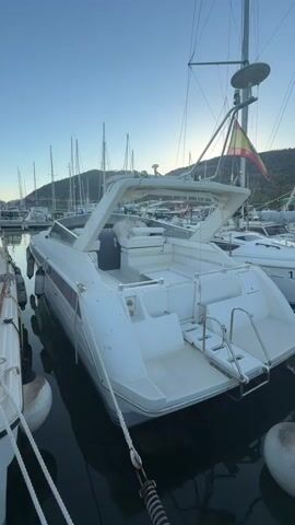 This media file captures a serene scene at a marina, likely in Sitges, Spain, bathed in the soft light of early morning or late evening. The primary subject is a white motor yacht, viewed from its stern. The boat features a white Bimini top providing shade over a seating area, with ample deck space visible. A retractable swim ladder is deployed, suggesting readiness for water activities. On the stern of the boat, the name "MWWE" is visible in dark lettering.

The boat is moored amongst other vessels, with numerous sailboat masts piercing the clear sky in the background. Hills or mountains rise in the distance, their slopes sparsely covered with vegetation. A Spanish flag flutters from a pole on the yacht, confirming the location's context. The water is calm, reflecting the boat and the surrounding marina, hinting at a peaceful day or the end of one.

The overall mood is tranquil and inviting, with the warm hues of the sky suggesting a pleasant atmosphere. The clear sky and gentle light indicate fair weather, ideal for boating. The presence of mooring lines and buoys further anchors the scene within a functioning marina environment. There are no people visible in the frame, emphasizing the quiet solitude of the moment. The focus is entirely on the boat and the peaceful marina setting.