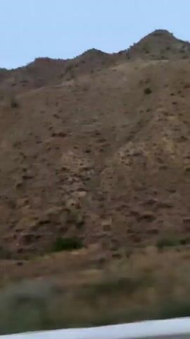 The video shows a landscape of rolling hills or mountains. The hills are covered in dry, brown grass and sparse vegetation. The sky is a pale blue, indicating it might be early morning or late evening. A guardrail with white and black stripes is visible in the foreground, suggesting the footage was captured from a moving vehicle on a road. There are no people or animals visible in the frame.