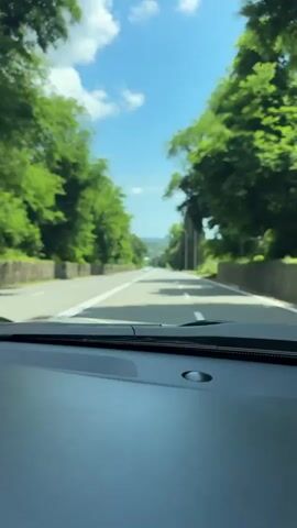 The media shows a first-person perspective from inside a car driving on a road. The road is paved with a white dividing line in the center and bordered by green trees and foliage on both sides. The sky is blue with scattered white clouds, suggesting daytime and pleasant weather. In the distance, the road curves slightly, and the landscape appears to be hilly or mountainous. There are a few cars visible on the road ahead, and some utility poles can be seen along the side. The dashboard of the car is visible in the foreground. The overall impression is that of a scenic drive through a natural environment.