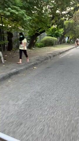 A first-person perspective video shows a person riding a bicycle down a paved road. The rider is wearing a light-colored shirt and dark pants. The road appears to be in an urban or suburban area, with trees and bushes lining the side. A wall made of grey blocks is visible behind the greenery. Other vehicles, including a motorcycle and a car, can be seen further down the road. The overall lighting suggests it is daytime, possibly late afternoon due to the warm glow.