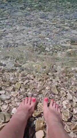 The video shows a close-up view of a person's feet partially submerged in clear, shallow water. The feet are on a rocky beach, with many smooth, light-colored stones visible beneath the water and on the shore. The water is calm, with gentle ripples moving across its surface. The toes are painted with bright pink nail polish. The sunlight appears to be bright, suggesting it is daytime. The overall scene evokes a sense of relaxation and enjoyment of a beach setting. No visible text is present in the video.