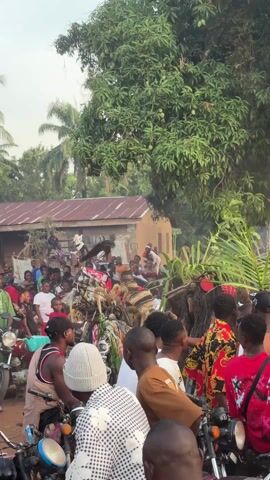 The video shows a crowd of people gathered around a building in a rural setting. Many of the people are wearing traditional African clothing and headdresses. Some of them are holding palm fronds. There are several motorcycles parked on the side of the road. In the background, there are palm trees and other vegetation.  The scene appears to be a cultural event or celebration.