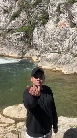 A person in a black sweatshirt and black baseball cap is standing in front of a body of water with a waterfall flowing into it. The person extends their hand towards the camera, then points to the waterfall. The waterfall is surrounded by steep, rocky cliffs. The water is clear and green, and the sky is partly cloudy.