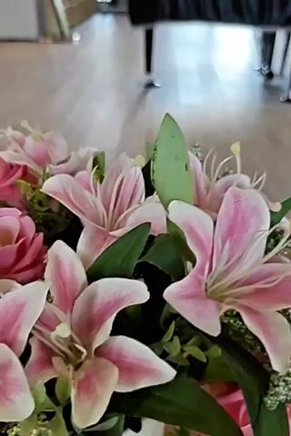 The video shows a close-up of a floral arrangement featuring pink lilies and roses. The flowers are the primary focus, with their petals and leaves clearly visible. The background is blurred, but appears to be an indoor setting with wooden flooring and furniture, possibly a room with a piano covered by a black cloth. The lighting suggests it is daytime. The camera movement is subtle, with a slight pan or tilt. There are no people or discernible actions occurring within the frame, other than the gentle movement of the camera.