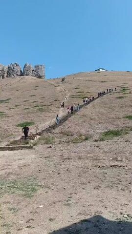The video shows a group of people climbing a long, steep staircase on a dry, grassy hillside. The staircase is made of stone or concrete and has a railing on one side. The people are spread out along the staircase, with some closer to the bottom and others further up. The hillside is covered in dry grass and some sparse vegetation. In the background, there are rocky outcrops and a clear blue sky. One person in a dark shirt is walking up the stairs in the foreground. The camera is positioned at the bottom of the staircase, looking up.