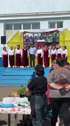 A wide, eye-level shot captures an outdoor performance. A mixed-gender choir of about a dozen elderly individuals is standing on a bright blue stage. They are dressed in similar outfits, mainly white shirts and maroon robes. A man is standing among them. A backdrop behind the stage includes yellow and white draping and a photo montage featuring images of people and the Russian flag.

In the foreground, two women are standing with their backs to the camera. The woman on the right is wearing a gray t-shirt with the words "DREAM TEAM" and an image of a dog. She holds a cell phone. The woman on the left has on a black shirt, a black cap, and jeans. A table with food and miscellaneous items is set up to the left of the women.

The overall setting appears to be a community event. The scene is well-lit, suggesting a sunny day.