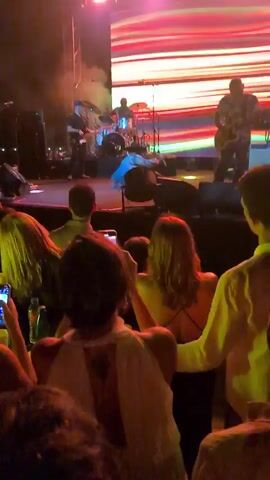 A low-angle, handheld video shows a live music performance. In the foreground, the backs of concertgoers' heads and shoulders are visible, some holding up phones to record. The audience is lit with green stage lights. The middle ground shows the stage itself, with band members playing instruments and a lead singer in a wide-brimmed hat and colorful, loose-fitting clothing dancing energetically. Behind the performers, a large LED screen displays a vibrant, abstract pattern of horizontal stripes in red, yellow, green, and white. Stage lights, including spotlights and overhead fixtures, illuminate the scene. Smoke or haze is visible on stage, adding to the atmosphere. A drum kit, microphones, and musical equipment are present on stage. The overall environment appears to be an outdoor or large indoor venue at night.