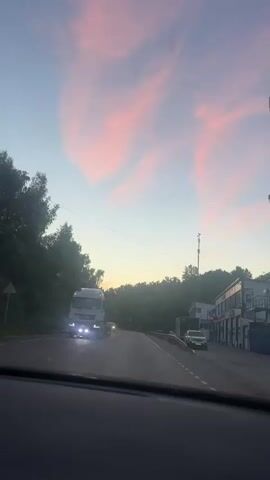The media file shows a view from inside a vehicle traveling on a road in Meshcherino, Russia. The time of day appears to be sunset or twilight, as indicated by the soft, diffused light and the vibrant pink and blue hues in the sky. Wispy clouds are scattered across the sky, catching the last rays of the sun.

The road curves gently to the right, bordered by a metal guardrail on the right side. On the left, a dense line of trees creates a natural backdrop, their dark foliage contrasting with the lighter sky. On the right side of the road, there is a building with several bays, suggesting it might be a garage, workshop, or commercial establishment. A cell tower is also visible next to the building.

Several vehicles are present. A large white truck with its headlights on is in the left lane, approaching the viewer. Further ahead on the same lane, a smaller silver car is visible. On the right side of the road, parked near the building, are a few other vehicles, including a dark sedan and a white van or small truck.

The overall mood is serene and peaceful, evoking the quiet beauty of a rural landscape at dusk. The scene captures a moment of transition, as day gives way to night, with the natural beauty of the sky and the subtle presence of human activity on the road. There is no discernible text within the frames.