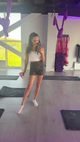 The image shows a woman taking a selfie in what appears to be a yoga or aerial fitness studio. The woman is wearing a white crop top, leopard print shorts, and white socks. She is holding a pink water bottle in her left hand. The studio has a wooden floor and a large window that lets in natural light. There are yoga mats on the floor, and purple aerial silks are hanging from the ceiling. In the background, there are stacks of purple yoga blocks. The overall impression is that the woman is at the gym, ready for a workout. The time of day appears to be daytime, based on the natural light coming through the window. The location is suggested to be Petropavlovsk-Kamchatsky, Russia. No visible text is present in the image.