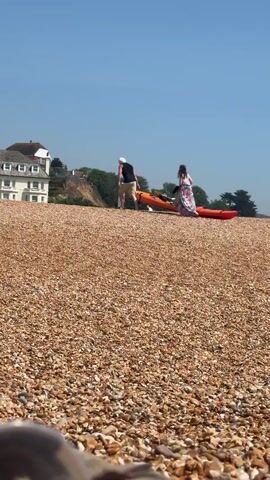 On a pebbly beach, a man and a woman are carrying kayaks. The man is on the left, wearing a black tank top and khaki shorts with a white hat. The woman is on the right wearing a floral dress. They are carrying an orange kayak and a red kayak. In the background, there is a large house and trees. The sky is blue.