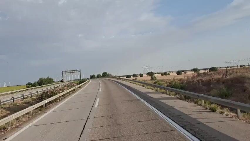 The video captures a view from a vehicle moving along a two-lane road. On the left side of the road, there is a barrier with flowers growing alongside it. Further down the road, a row of tall structures, possibly for power lines, is visible. The right side of the road features a metal guardrail. Beyond the road and guardrail, the landscape consists of dry, yellowish fields with scattered trees and a distant row of wind turbines. The sky is partly cloudy. The camera perspective suggests the video is taken from the driver's or front passenger's seat of the moving vehicle.