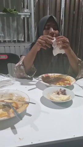 A person wearing a gray hijab and a patterned top sits at a white table, using a white napkin to wipe their face. In front of them are two plates with leftover food, and a small bowl containing food and pieces of fruit. The setting appears to be outdoors, possibly a restaurant or an open-air dining area, with a wooden fence and curtains visible in the background. The lighting suggests it is nighttime or evening. The person's actions with the napkin might indicate they are wiping away sweat, tears, or simply cleaning their face after eating. No visible text is present in the frame.