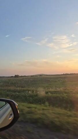 This is a vertical, first-person perspective video filmed from a moving vehicle. The camera is positioned to capture the view out of the side of the vehicle, including the side mirror which is visible in the lower left corner of the frame. The scene is an open field with tall grass and some wildflowers, likely a rural landscape. In the distance, there are trees and a gently rolling hill. The sky is a clear blue with a few wispy clouds, and the sun is setting on the right side of the frame, casting a warm, orange glow over the landscape. The road the vehicle is traveling on is visible in the lower part of the frame, curving towards the right. There are no people visible in the video. The overall mood is peaceful and serene, capturing the beauty of a sunset in a rural setting.