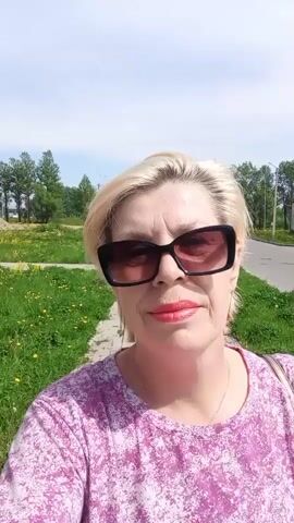 A blonde woman wearing sunglasses and a pink floral top is taking a selfie video. She is standing on a sidewalk next to a grassy area dotted with yellow flowers. In the background, there are trees, streetlights, and buildings. The sky is blue with some clouds.  A vehicle is briefly visible passing in the background.