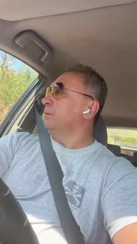 A man in his late 40s or early 50s with short gray hair and sunglasses is driving a car. He is wearing a gray t-shirt with a skull and crossbones graphic. He has a seatbelt on and is wearing white wireless earbuds. The car is moving along a road through a dry, hilly landscape. The sun is shining.