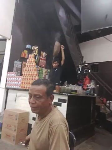 The video shows the interior of a small store with an upstairs loft. A man in a tan shirt stands in the foreground and looks at the camera while smoking a cigarette. Behind him, a younger man in a black shirt is preparing something behind a counter laden with stacked goods. Another man, in a red and blue plaid shirt and a Muslim prayer cap, stands behind the counter. The background is dark, with stairs leading to the loft and a TV mounted on the wall.