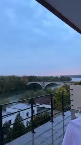 From a high balcony overlooking a river, the camera pans slightly to the right, revealing a grey, overcast sky and a landscape featuring a river with treed islands and a stone bridge. Several modern, low-rise buildings can be seen closer to the viewpoint, behind a black metal railing. In the far distance, the silhouette of a city skyline is visible. The balcony floor appears to be damp, suggesting recent rain. The overall tone is muted and calm. The camera remains static for the duration of the short clip.