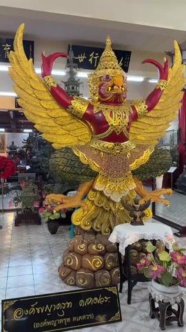 The video shows a large, ornate statue of a Garuda, a mythical bird-like creature, with bright red and gold coloring. The statue is positioned in the center of a room, with its wings spread wide. The Garuda has a bird-like head with a beak and eyes, and it wears an elaborate headdress and jewelry. Its body is adorned with intricate patterns and gold leaf. It has multiple arms and hands, with sharp talons. The statue is mounted on a large, cloud-like base with decorative elements. In front of the statue, there is a small table covered with a white cloth, displaying a smaller statue of a bird, and a pot with pink and purple flowers. There is also a sign with Thai writing in front of the statue. The background of the room reveals more statues, some of which appear to be figures in traditional attire, and Chinese calligraphy written on scrolls hanging on the wall. A traditional Chinese lantern is also visible. The floor is tiled. The overall impression is that of a display within a temple or a museum dedicated to religious or cultural artifacts.