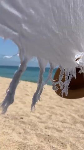 This media file captures a serene beach scene at Sant Pol de Mar, Spain. The foreground is dominated by a white beach towel or fabric, partially obscuring a straw hat and a wooden pole, suggesting an umbrella setup. The fabric has a frayed, tasseled edge, with intricate white cords twisted together. The hat, made of woven straw, is visible beneath the fabric, casting a subtle shadow.

The background opens up to a tranquil beach with fine, light-colored sand. Beyond the sand lies the glistening turquoise sea, meeting a clear blue sky dotted with wispy white clouds. The horizon is distinctly visible, creating a sense of vastness and peace. A faint yellow marker or buoy can be seen in the distance on the water.

The time of day appears to be daytime, likely late morning or early afternoon, given the bright sunlight and the clear blue sky. The weather seems warm and pleasant, ideal for a beach outing. The overall mood is one of relaxation and tranquility, evoking the feeling of a peaceful day at the beach. There is no visible text in this media file.