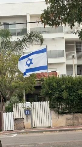 The video shows an Israeli flag waving in front of a two-story residential building with a white fence and the number 11 on the gate. There are trees on either side of the flag, and the sky is overcast. A voice says something in another language.