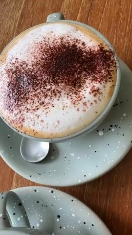 The video shows a table at a cafe, laden with various sweet treats and coffees. The scene begins with a close-up of a cappuccino dusted with cocoa powder, resting on a pale green speckled saucer with a spoon. The camera then pans to reveal a second, larger cup of coffee with latte art, also on a similar saucer. Further to the right, a slice of tiramisu is presented on a white plate, garnished with a sprig of rosemary. The camera then shifts to show a slice of lemon tart with meringue and blueberries on another white plate. The table is made of wooden planks, and the overall atmosphere is cozy and inviting, suggesting a delightful cafe experience.