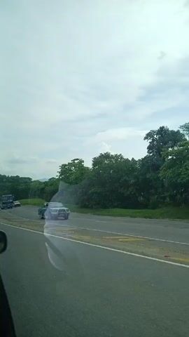The video shows a view from a moving vehicle, likely a car, driving on a paved highway. Initially, a teal-colored pickup truck with a bull bar is visible on the opposite side of the road, moving in the same direction. Then, a large blue bus labeled "INTER AFRICA" passes on the opposite side. The landscape features green trees and grassy verges beside the road. In the distance, there are hills or low mountains under a cloudy sky. A metal safety barrier is briefly visible alongside the road. The overall scene suggests a rural or semi-rural environment with vehicles traveling on a highway.