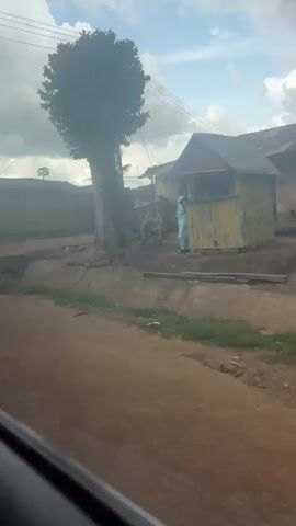 The video shows a moving shot of a dirt road with several buildings and people. The sky is cloudy. On the left side of the road, there is a ditch with a small bridge. Further down the road, there are buildings made of concrete and corrugated metal. Several people are walking or sitting outside the buildings. One person is wearing a dark headscarf and a long dress, and is walking away from the camera. Another person, a man in a light-colored outfit, is walking towards the camera, carrying a dark object in his right hand. There is a building with a white facade and a gray base, featuring an arched entrance. Further along, there is a building with two large black water tanks on the roof. Another building appears to be a shop with a sign that says "Tomar's". People are seen standing outside this shop. The overall scene depicts a residential or commercial area in what appears to be a developing country.