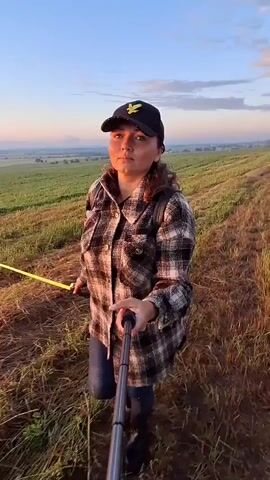 The video shows a person holding a selfie stick and recording themselves in a field. The person is wearing a plaid shirt, jeans, and a baseball cap. The field appears to be covered in dry grass and there is a gentle slope in the background. The sky is a soft blue with some clouds, suggesting it might be sunrise or sunset. The person is looking at the camera and appears to be walking or moving slightly, as indicated by the slight blur in some frames. The overall atmosphere is calm and serene.