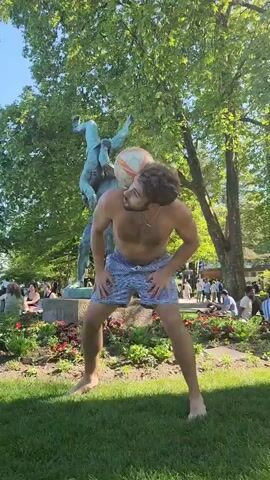 A shirtless man in blue and white striped shorts juggles a soccer ball in a park. He is barefoot on the grass, using his head, chest, and knees to keep the ball in the air. Behind him is a bronze statue of two people intertwined, and a flower bed with red and white flowers.  In the background, people are sitting and standing around the park. There are trees providing shade and a clear blue sky above.